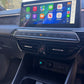 A car interior dashboard with a large touchscreen display showing Apple CarPlay interface, mounted above climate controls and a center console storage compartment.